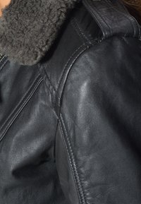 Zwarte leren jas met een grijze schapenvacht kraag, met zichtbare stiksels en een gladde textuur. Close-up van het schoudergedeelte.