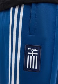 Μπλε αθλητικό σορτς με λευκές κάθετες ρίγες και ένα σήμα με ελληνικό κείμενο και σχέδιο σταυρού κοντά στην τσέπη.