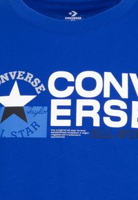 Camiseta de algodón azul con texto grande en blanco, un gráfico de estrella y detalles de marca. Incluye información impresa sobre la marca y el logo.