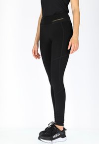 Zwarte leggings met een hoge tailleband, voorzien van subtiele stiksel details en een gouden logo. Gedragen met zwarte sportschoenen op een witte achtergrond.