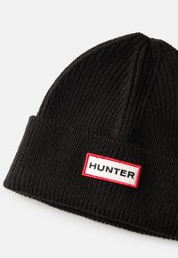 Černá pletená čepice s ribbovanou texturou, která má obdélníkovou bílou etiketu orámovanou červenou barvou se slovem "HUNTER" černým písmem.