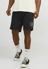 Des shorts en coton noirs présentent un logo sur la jambe gauche, associés à des chaussettes blanches et des baskets noires avec des lacets blancs sur un fond neutre.