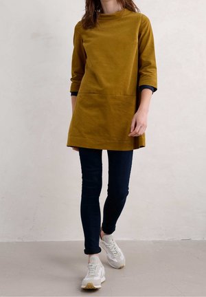 Sennepsgul tunika kjole med høj halsudskæring, 3/4-ærmer og frontlomme. Parret med mørke jeans og lyse sneakers.