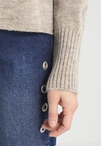 Hand, die in der Nähe eines blauen Jeansrocks mit Metallösen ruht, trägt einen beigefarbenen, gerippten Strickpullover.