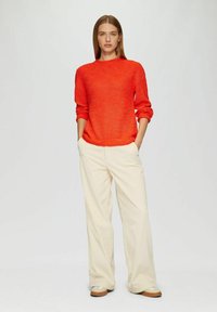 Leuchtend roter Strickpullover mit rundem Ausschnitt, kombiniert mit cremfarbenen weit geschnittenen Hosen und hellbraunen Slippern. Sauberes und schlichtes Design.