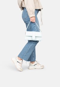 Borsa azzurro chiaro con manico superiore, forma rettangolare, finitura liscia e chiusura con patta. Abbinata a jeans di jeans blu e sneakers bianche.