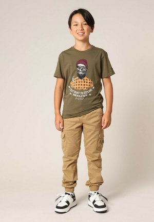 T-shirt vert olive avec un imprimé graphique, associé à un pantalon cargo marron clair. Baskets noires et blanches avec lacets. Tenue simple et décontractée.