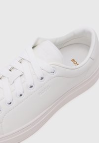 Sneaker bianco con tomaia in pelle liscia, punta tonda, lacci piatti, logo del marchio impresso e design minimalista. Suola in gomma e interni imbottiti.
