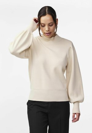 Vrouw met donker haar draagt een crèmekleurige trui met pofmouwen en zwarte broek, kijkt omlaag met één hand die haar hoofd aanraakt.