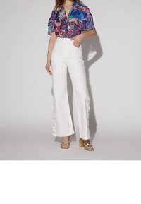 Blouse multicolore à motifs floraux avec des manches courtes volantées, assortie à un pantalon large blanc taille haute avec des boutons décoratifs sur le côté.