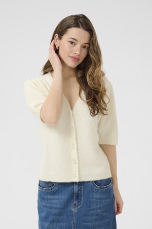 Mlada žena s dugom valovitom kosom, odjevena u kremasti kratki kardigan s gumbima i plavu traper suknju, dodiruje uho jednom rukom.