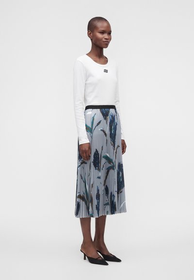 Femme portant un haut blanc à manches longues et une jupe midi plissée avec un motif floral bleu et vert, talons slingback noirs, debout devant un fond blanc.