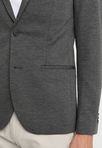 Blazer grigio scuro a un bottone indossato con pantaloni chiari, mostra la tasca frontale e il braccio con dettagli a bottone sulla manica.