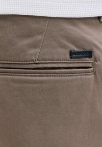 Gros plan sur la poche arrière beige d'un pantalon avec une étiquette noire Jack & Jones cousue au-dessus de la couture de la poche, portée avec une chemise blanche texturée.