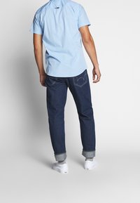 Ljusblå kortärmad skjorta i kombination med mörka jeans med uppvikta ärmar och vita sneakers, sedd bakifrån.