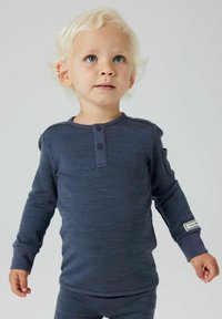 Kleinkind mit lockigem blonden Haar, das ein navyblaues Langarmhemd und eine Hose trägt, das leicht nach oben schaut, vor einem schlichten Hintergrund.