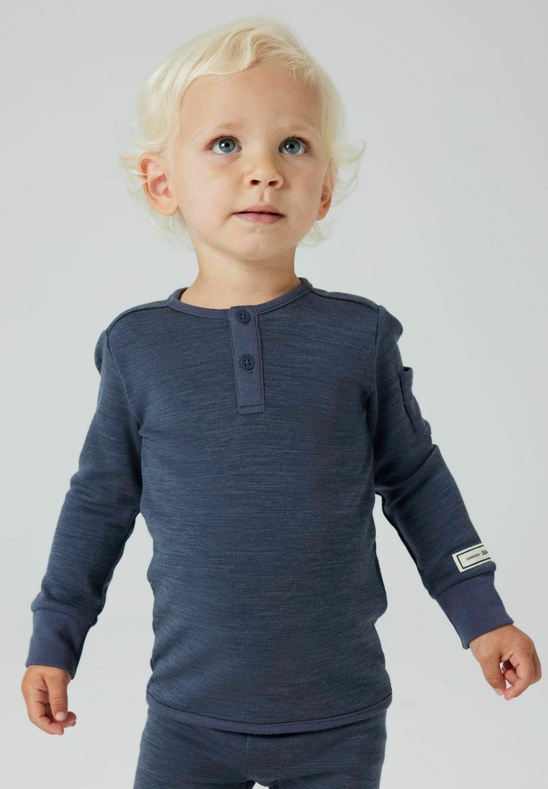 Kleinkind mit lockigem blonden Haar, das ein navyblaues Langarmhemd und eine Hose trägt, das leicht nach oben schaut, vor einem schlichten Hintergrund.