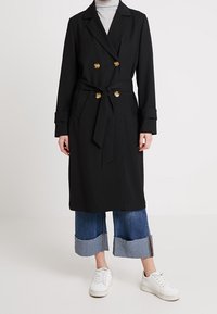 Svart dubbelknäppt trenchcoat med guldknappar och midjeband, bärs över uppvikta blå jeans och vita sneakers.