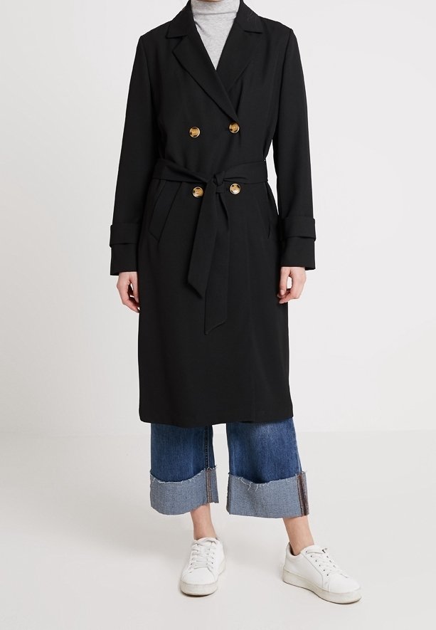 Svart dubbelknäppt trenchcoat med guldknappar och midjeband, bärs över uppvikta blå jeans och vita sneakers.