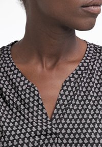 Nahaufnahme des Nackens und des unteren Gesichts einer Person mit dunkler Haut, die ein schwarzes Oberteil mit kleinen weißen Mustern trägt.