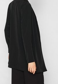 Haut noir à manches longues, en tissu doux, avec une coupe décontractée, un ourlet arrière plus long, un design minimaliste et sans détails apparents en métal.
