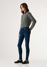 Suéter gris de punto con patrón texturizado, combinado con vaqueros ajustados azul oscuro. La modelo lleva zapatos negros sin cordones, posando sobre un fondo neutro.