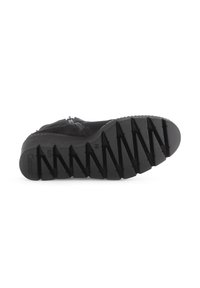 Zwarte schoen met een textured rubberen zool, voorzien van diagonale groeven voor grip, een korte rits en een zacht bovenmateriaal.