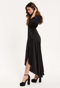 Black satin maxi dress with a fitted bodice, puff sleeves, and side slit; styled with black platform heels and ankle strap detail.