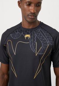 Hombre con camiseta atlética negra con logotipo de colmillo de serpiente dorado y patrón abstracto de hojas grises en el pecho, mirando hacia abajo.