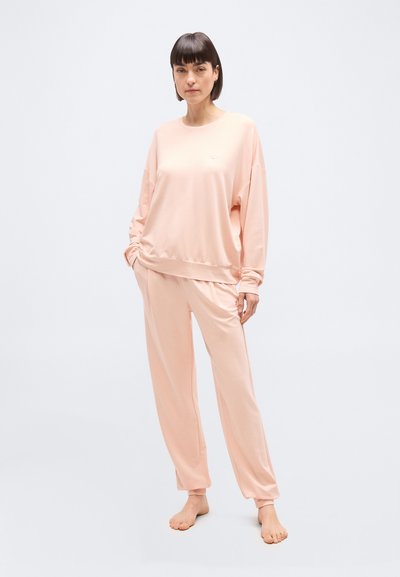 Γυναίκα που στέκεται ξυπόλυτη φορώντας χαλαρό, ανοιχτό ροδακινί μπλουζάκι με μακριά μανίκια και παντελόνι που ταιριάζει, πάνω σε απλό λευκό φόντο.