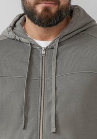 Grijze ritssluiting hoodie met een gevoerde capuchon, verstelbare trekkoorden en textiel met horizontale naaddetails. Zilveren ritssluiting.