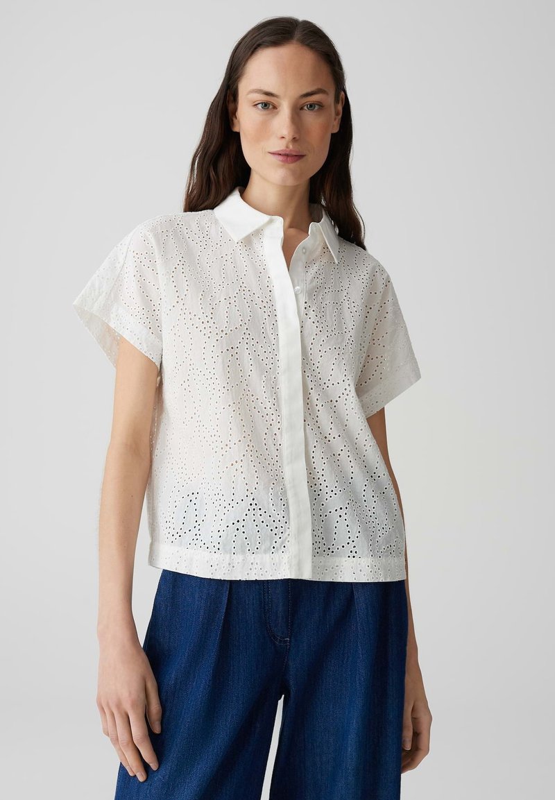 Camicetta bianca a maniche corte realizzata in tessuto leggero con fori tagliati a laser e chiusura con bottoni, abbinata a pantaloni larghi blu scuro.