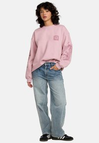 Sweatshirt roz cu mâneci lungi, prezentând imprimeuri grafice pe mâneci și piept, asortat cu blugi lejeri de culoare albastru deschis și adidași negri.