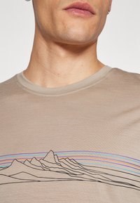 Beige T-shirt met een ronde hals, voorzien van een zwart lijntekening van bergen en kleurrijke horizontale strepen boven de afbeelding.
