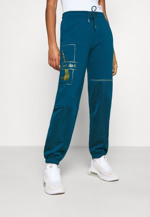 Osoba ubrana w turkusowe spodnie dresowe Nike ze złotym logo "Just do it." na lewej udzie oraz białe buty sportowe Nike z widocznymi podeszwami powietrznymi.