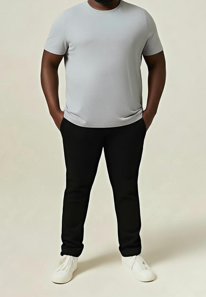 Homme portant un t-shirt à manches courtes gris clair, pantalon noir, baskets blanches, debout les mains dans les poches devant un fond clair.