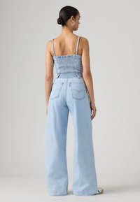 Γυναίκα με κότσο, φορά σκουλαρίκια κρίκους, αμάνικη μπλούζα από ελαφρύ τζιν με σφηκοφωλιά, και φαρδιά ανοιχτόχρωμα μπλε τζιν παντελόνια, στέκεται με την πλάτη προς τα εμπρός.