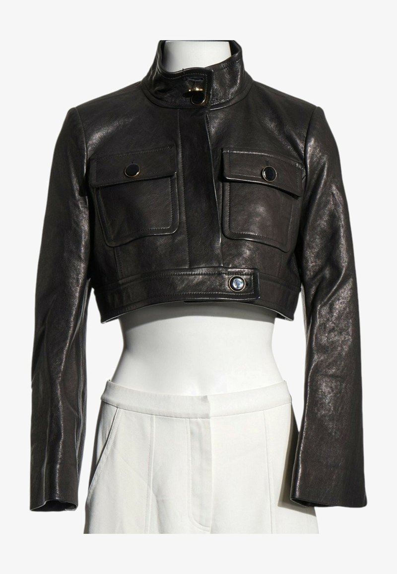 Veste en cuir noire courte avec manches longues, col montant, deux poches poitrine boutonnées et une ceinture boutonnée à la taille portée avec un pantalon blanc.