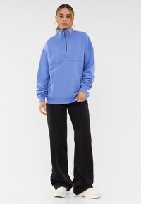Lichtblauwe half-zip pullover met een relaxte pasvorm, voorzien van een kangoeroezak en ribgebreide manchetten, gecombineerd met brede zwarte broeken en witte sneakers.