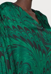 Grön och svart mönstrad blus med v-ringning, slät textur och knappdetalj nära kragen. Tyget faller elegant.