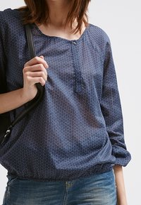 Tmavě modrá halenka s puntíky, kulatým výstřihem, knoflíkovou légou na dva knoflíky, raglánovými rukávy a elastickým lemem. Nosit s džíny a černou kabelkou.