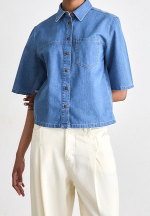 Person iført en kortærmet blå denim skjorte med knapper og forreste lomme samt off-white højtaljede bukser mod en ensfarvet baggrund.
