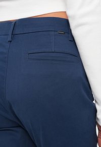 Marineblå bukser laget av glatt stoff, med beltehempe, baklomme med klaff og en liten merkevarelogo på midjebåndet.