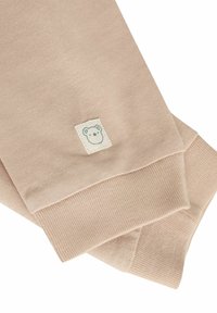 Jasno beżowy sweter z bawełny z prążkowanymi mankietami, zdobiony małą kwadratową metką z ilustracją misia. Gładka faktura, prosty design.