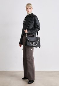 Borsa a tracolla nera in pelle con accentuazione in catena argentata, forma rettangolare e chiusura a pattina. Il modello indossa una giacca di pelle nera e pantaloni marroni.