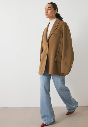 Blazer oversize in misto lana marrone con un solo bottone, tasche a toppa, abbinato a jeans a gamba larga blu chiaro e décolleté rosse a punta.