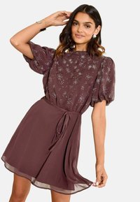 Rochie maro cu mâneci scurte tip puff, ornamente cu model de stele, talie strânsă și fustă fluidă. Material ușor, moale, cu o textură netedă.