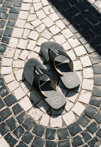 Γκρίζες σαγιονάρες Havaianas που ακουμπούν πάνω σε ασπρόμαυρο ψηφιδωτό πλακόστρωτο υπό το φως και τη σκιά του ήλιου.