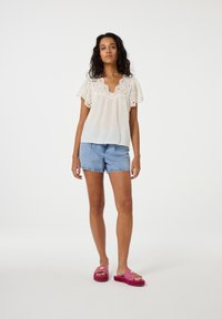 Witte korte mouwen top met oogjesdetails, gecombineerd met blauwe denim shorts. Schoeisel bestaat uit roze slides met bloemaccenten.