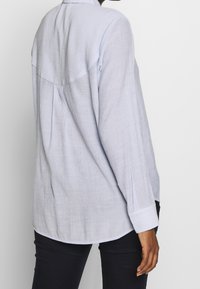 Camicia azzurra a maniche lunghe con tessuto testurizzato, orlo arrotondato, pieghe sul retro e polsini con bottoni. Abbinata a pantaloni neri.